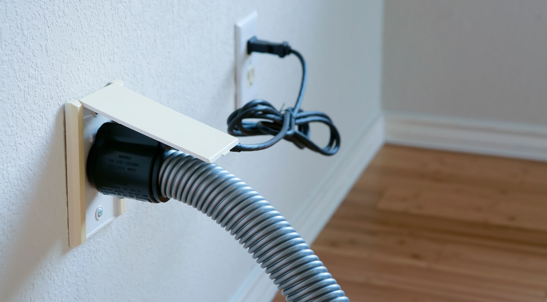 Central vacuum hose plugged into a wall inlet with power cord and hardwood floor in the background.